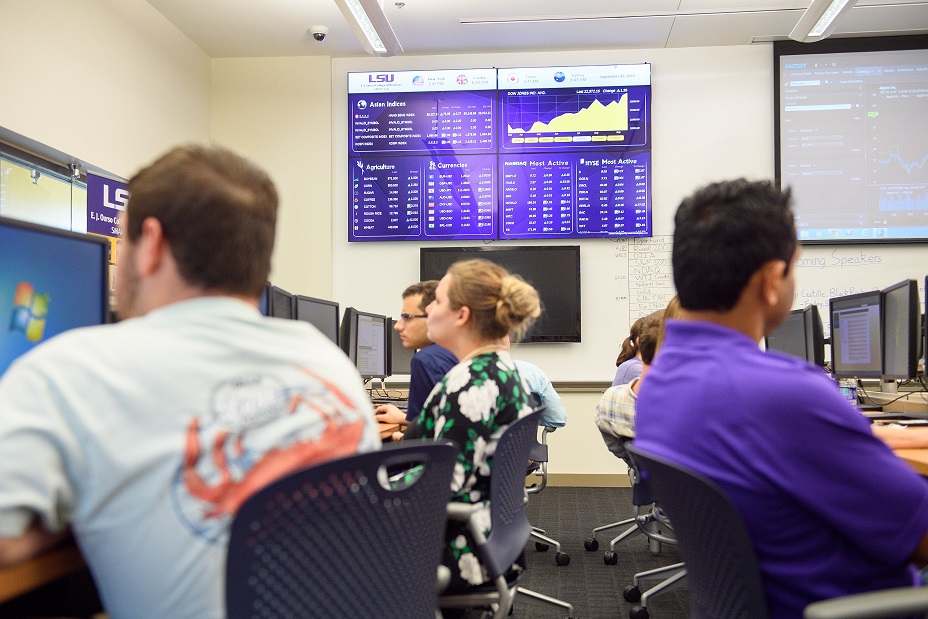 Students sitting a computers. A screen in the background displays financial data.